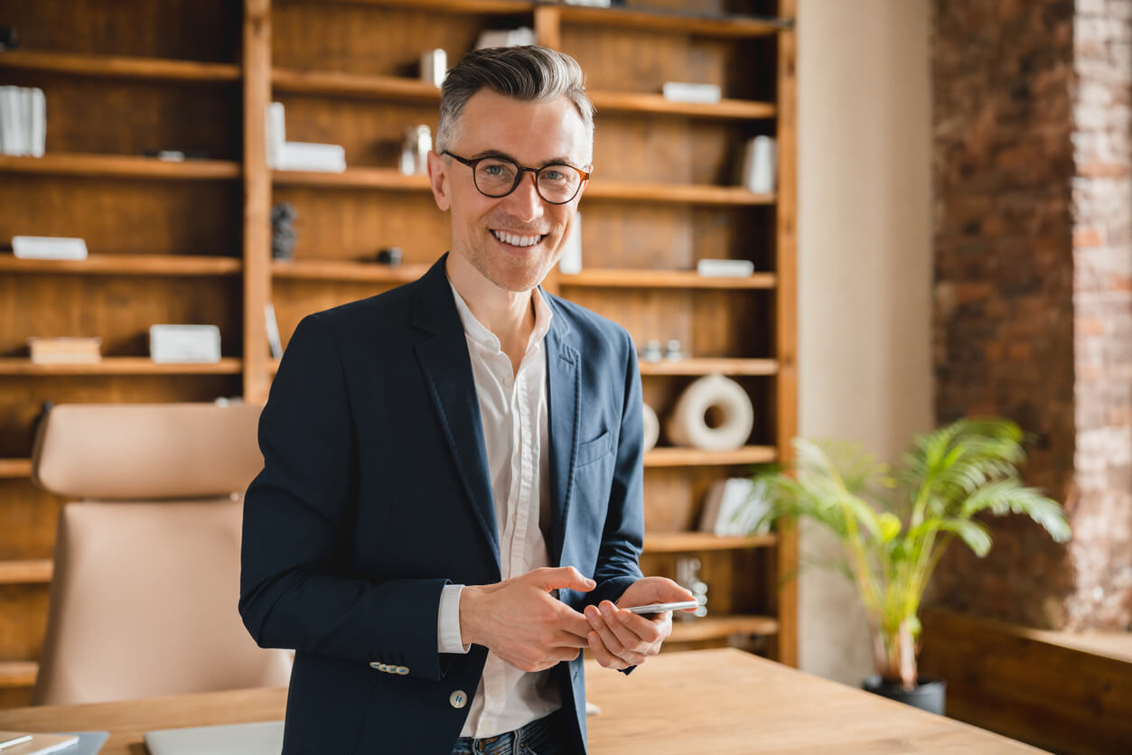 Un homme d'affaires souriant, tenant un smartphone, se tient dans un bureau élégant. En arrière-plan, étagères en bois, fournitures ordonnées et plante décorative.