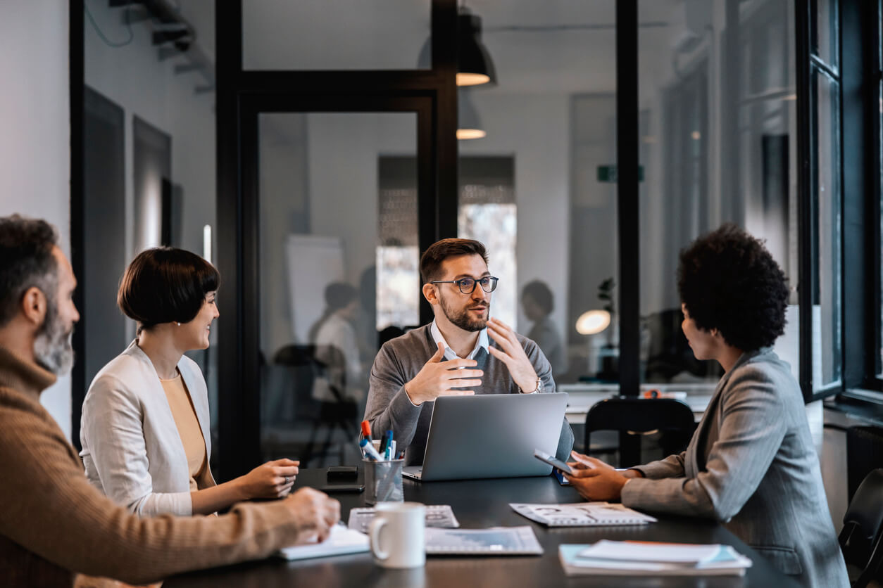 Un groupe de professionnels discute autour d'une table dans une salle de réunion moderne. L'un d'eux, au centre, explique un point avec des gestes, un ordinateur portable devant lui. Cette scène illustre une collaboration stratégique nécessaire pour la gestion d'une société civile immobilière