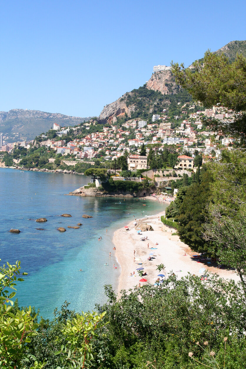 Une plage de la Côte d'Azur, bordée par des eaux turquoise, avec en arrière-plan la ville de Beausoleil nichée à flanc de colline. On y voit des maisons colorées surplombant la mer, entourées de végétation méditerranéenne et de falaises rocheuses. Des vacanciers sont installés sur la plage de galets avec des parasols colorés.