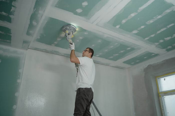 Un homme en train de rénover la toiture d'une maison. Il se tient sur une échelle. On voit autour de lui que le plafond et les murs ont été rénovés.