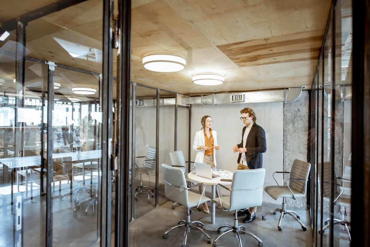 L'image montre un local d'entreprise moderne avec des bureaux équipés, ordinateurs et documents. Un homme et une femme échangent sur les enjeux fiscaux et comptables d'une SCI pour entreprises, mettant en avant l'importance d'une gestion stratégique de l'immobilier d'entreprise.