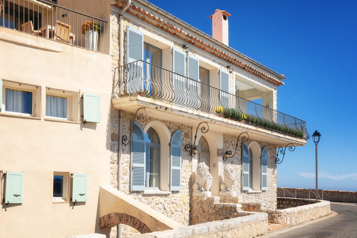 Façade en pierre d'une villa méditerranéenne avec des volets bleus et une large terrasse, décorée de deux statues de lions. La villa est située à Antibes, face à un ciel bleu dégagé. Elle possède des détails architecturaux élégants, comme des balustrades en fer forgé et des volets aux tons pastel