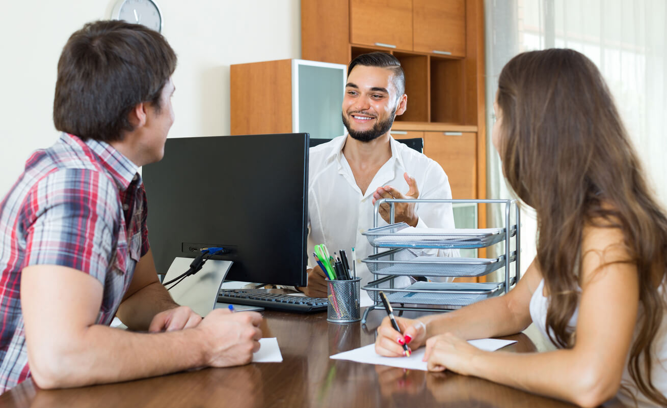 Trois personnes discutent dans un bureau équipé d'ordinateurs, examinant des documents et échangeant sur les étapes pour déclarer un statut LMNP en ligne de manière efficace.