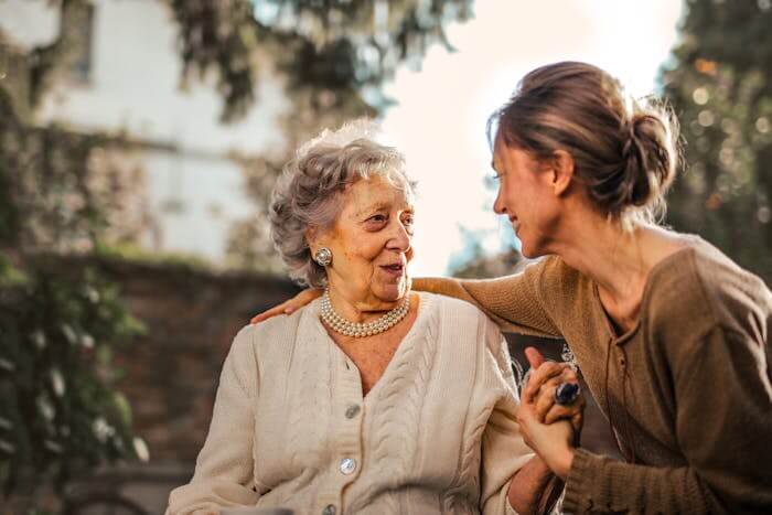 Une jeune femme souriante rendant visite à sa mère dans une résidence senior, les deux se tenant par la main dans le jardin